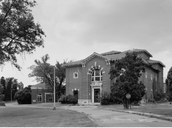 A black and white image of a two-story brick structure surrounded by deciduous trees. A road leads up to the building.