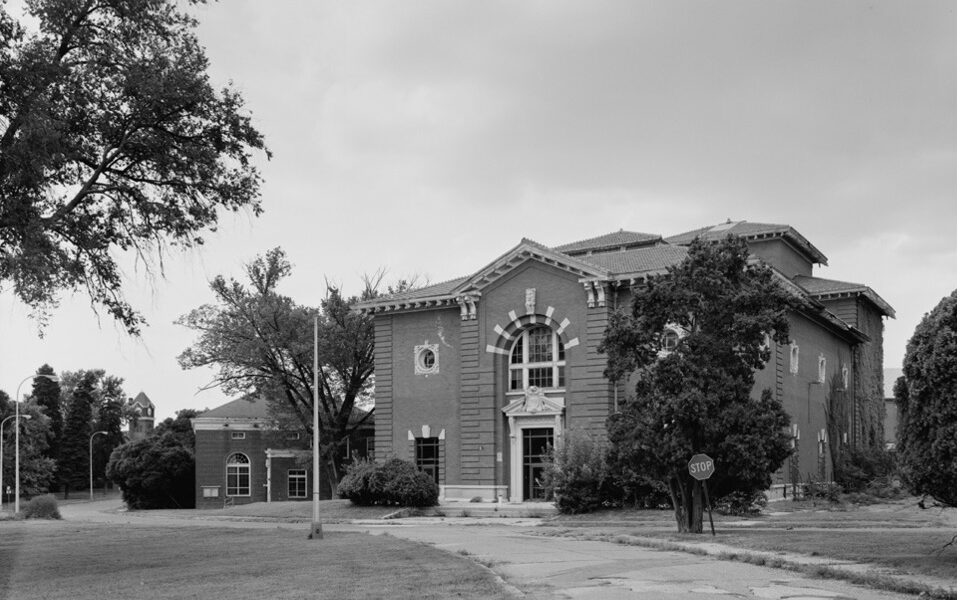 A black and white image of a two-story brick structure surrounded by deciduous trees. A road leads up to the building.