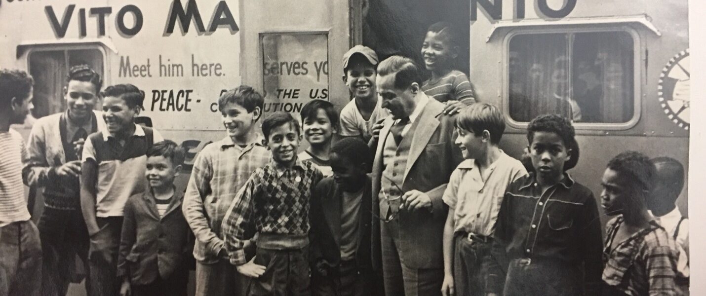 Rep. Marcantonio at his office, a trailer, with neighborhood children, ranging in age, from very young to teenagers. They are standing, some posing, some talking, with Marcantonio. The door to the trailer is open with some children standing inside the door frame, laughing. Marcantonio looks down at the child to his right, smiling warmly, with their arms around each other. The trailer reads "Congressional Office of your...Congressman," "Vito Ma....nio, "Meet him here," "PEACE," "Serves you..." Ellipsis in text signals what is blocked by the open door. Buildings line the top frame of the photo, behind Marcantonio's office.