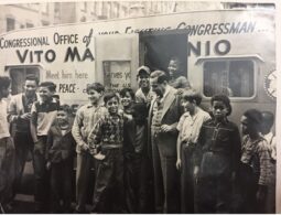 Rep. Marcantonio at his office, a trailer, with neighborhood children, ranging in age, from very young to teenagers. They are standing, some posing, some talking, with Marcantonio. The door to the trailer is open with some children standing inside the door frame, laughing. Marcantonio looks down at the child to his right, smiling warmly, with their arms around each other. The trailer reads "Congressional Office of your...Congressman," "Vito Ma....nio, "Meet him here," "PEACE," "Serves you..." Ellipsis in text signals what is blocked by the open door. Buildings line the top frame of the photo, behind Marcantonio's office.