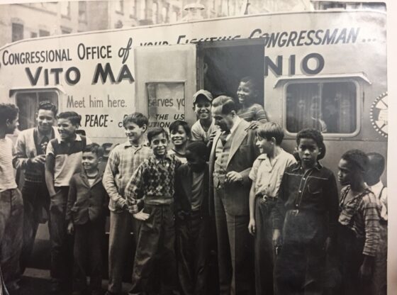 Rep. Marcantonio at his office, a trailer, with neighborhood children, ranging in age, from very young to teenagers. They are standing, some posing, some talking, with Marcantonio. The door to the trailer is open with some children standing inside the door frame, laughing. Marcantonio looks down at the child to his right, smiling warmly, with their arms around each other. The trailer reads "Congressional Office of your...Congressman," "Vito Ma....nio, "Meet him here," "PEACE," "Serves you..." Ellipsis in text signals what is blocked by the open door. Buildings line the top frame of the photo, behind Marcantonio's office.