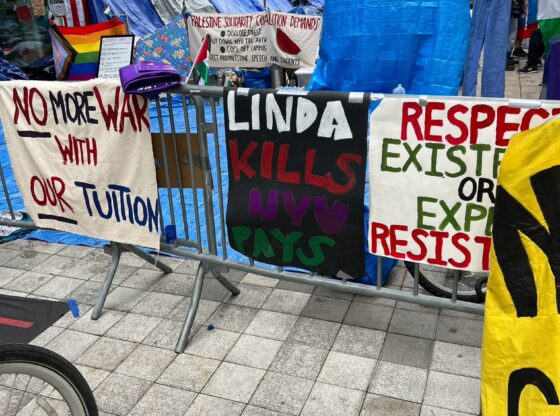 A picture of signs outside metal stand-up gates. The signs read: "Linda Kills / NYU Pays," "No More War with OUR Tuition," and other signs that are only partially in the frame. Behind the signs and gates are the tents that form the encampment. New York buildings are behind them.