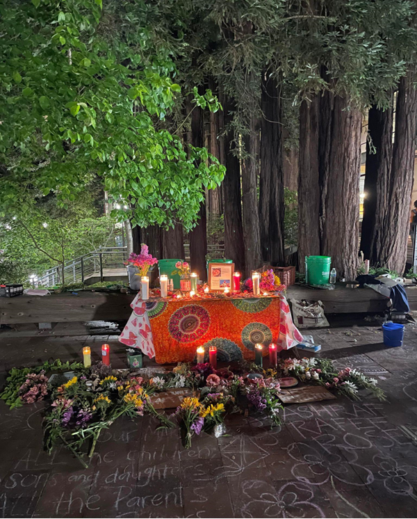 A memorial alter on top of a table, covered in vibrant fabrics, in orange, pink and other colors. There are memorial candles and flowers on top of the table and around it. Redwood tree trunks appear in the background and foliage from a tree out of frame peers into the top left corner. 