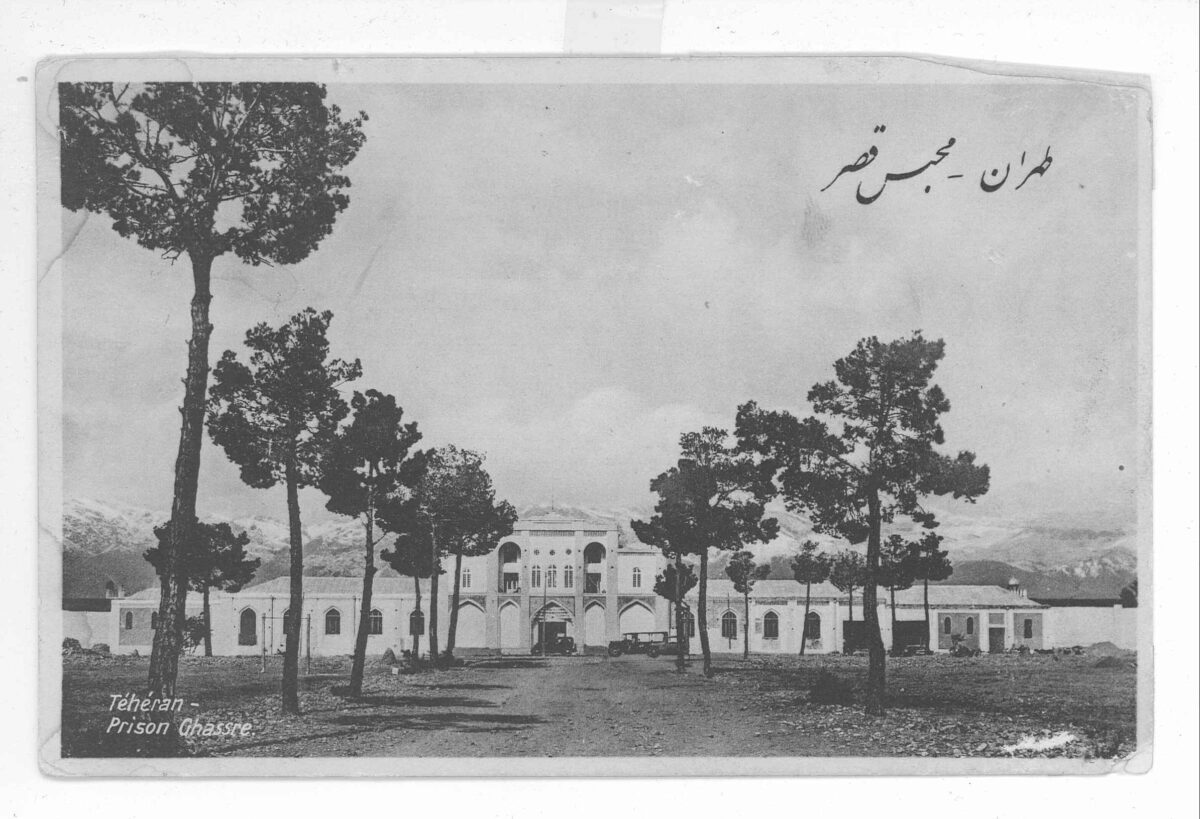 A black and white image of a large building - a prison, actually - is pictured in the distance, shot from a tree-lined road leading toward it. 