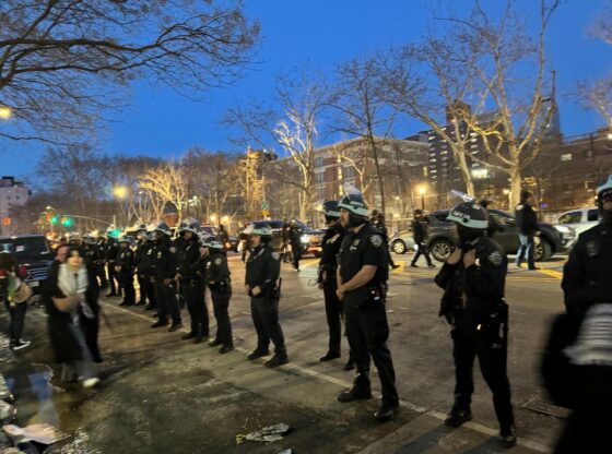 A color photograph depicts a line of NYPD amassing to crack down on the International Women's Day Global Strike for Gaza. They are standing along the shoulder of a city street as passerby walk in front of them. A chain link fence can be seen in the background with several city buildings behind it during an early evening.